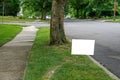 A white blank advertisement sign on a grassy strip near a street and a tree trunk Royalty Free Stock Photo