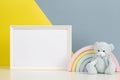 White blank frame with teddy bear and kid toy rainbow on white desk and yellow and gray background