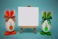 White blank paper on easel with funny egg in form of cute bunny on blue background. Happy easter.