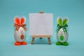 White blank paper on easel with funny eggs in form of cute bunny on blue background