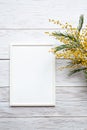 White blank photo frame with a bouquet of mimosa on a white wooden table. Minimalistic, mockup.