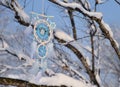 White and blue dreamcatcher hanging on tree branches covered with snow.