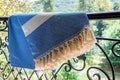 A white and blue Turkish peshtemal / towel on a wrought iron railings with blurry nature in the background.