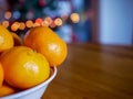White bowl filled with delicious clementine`s on pine wood table, glowing Christmas tree in the background Royalty Free Stock Photo