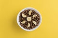 White bowl full of chocolate cereal balls and banana slices on yellow background Royalty Free Stock Photo