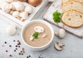 White bowl plate of creamy chestnut champignon mushroom soup on white kitchen background and box of raw mushrooms and fresh bread Royalty Free Stock Photo