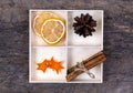 A white box with compartments on a wooden background filled with sticks of cinnamon, dried oranges, tangerine stars and tree cone