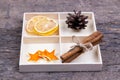 A white box with compartments on a wooden background filled with sticks of cinnamon, dried oranges, tangerine stars and tree cone