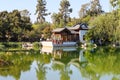 A white and brown Chinese pavilion in the garden with people walking through the pavilion near a deep green lake with lush green t Royalty Free Stock Photo