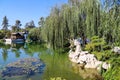 A white and brown Chinese pavilion in the garden with people walking through the pavilion near a deep green lake with lush green t Royalty Free Stock Photo