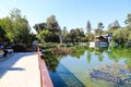 A white and brown Chinese pavilion in the garden with people walking through the pavilion near a deep green lake with lush green t Royalty Free Stock Photo