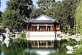 A white and brown Chinese pavilion in the garden with people walking through the pavilion near a deep green lake with lush green t Royalty Free Stock Photo