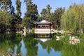 A white and brown Chinese pavilion in the garden with people walking through the pavilion near a deep green lake with lush green t Royalty Free Stock Photo