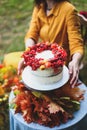 White cake with berries, maple leaves, apples, rowan. Woman in yellow dress on wooden chair by round table with blue tablecloth, Royalty Free Stock Photo