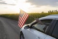 car with an American flag hanging from the window drives along country road among rapeseed field towards the setting sun Royalty Free Stock Photo