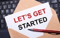White card with the text LET IS GET STARTED in a craft envelope on a work desk with a modern laptop keyboard and burgundy pen. Royalty Free Stock Photo