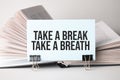 A white card with the text Take a break,take a breath stands on a clip for papers on the table against the background of books. Royalty Free Stock Photo