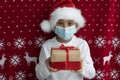White caucasian boy in santas hat and face medical mask holds two gift boxes tired with ribbons on red christmas background.