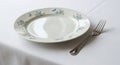 White ceramic plate with light blue floral patterns is set on a white tablecloth. A stainless steel Royalty Free Stock Photo