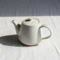 A white ceramic teapot is centered on a white, textured tablecloth, casting a soft Royalty Free Stock Photo