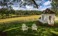 2 white chairs sit on a lawn in front of a white fence under a broad tree.