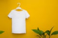 White children`s t-shirt hanging on a yellow background next to green leaves. Mocap. Copyspace