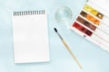 White clean note pad on black spring, squirrel brush, jar of water and watercolor paints on blue paper background Royalty Free Stock Photo