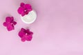 White container with cream for face and body with three magenta colored orchid flowers on pink background