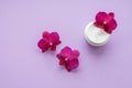 White container with cream for face and body with three magenta colored orchid flowers on purple background. Concept