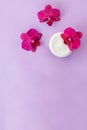 White container with cream for face and body with three magenta colored orchid flowers on purple background. Concept
