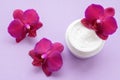 White container with cream for face and body with three magenta colored orchid flowers on purple background.
