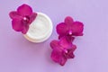 White container with cream for face and body with three magenta colored orchid flowers on purple background
