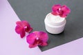 White container with cream for face and body with three magenta colored orchid flowers on purple and black background. Concept Royalty Free Stock Photo