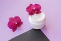 White container with cream for face and body with two magenta colored orchid flowers on purple and black background. Concept of de Royalty Free Stock Photo