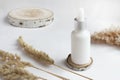 A white cosmetic bottle stands on a wooden stand on a white table, next to dried flowers. Serum, spa treatments, natural cosmetics