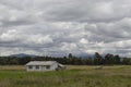 White country house with yellow green meadow, green trees, blue mountains and grey sky at background