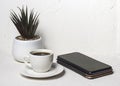 White cup with black aromatic coffee on a white abstract background with a potted flower in the background with a smartphone Royalty Free Stock Photo