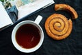 Cup of tea next to a cinnamon bun and an open book on a dark background Royalty Free Stock Photo