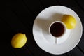 A white cup with black tea stands in a saucer on a dark wooden table. Nearby are two yellow lemons. View from above. Minimalism