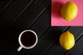 A white cup with black tea stands on a black table next to a pink napkin and two yellow lemons. View from above. Minimalism