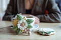 White cup of coffee on saucer and nice icing flower shaped cookies on table on foreground closeup, silhouette of lady on