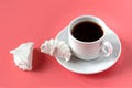 A white cup with coffee on a saucer and a white vanilla marshmallow broken into two halves on a pink background