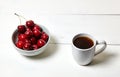 White cup with coffee and sweet cherries in a ceramic bowl on a white wooden background, closeup Royalty Free Stock Photo