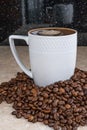 A white cup filled with roasted cinnamon coffee beans standing on brown marble surrounded by coffee beans