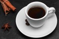 White cup of fresh hot black coffee, several coffee beans on a saucer, cinnamon sticks, anise star on black background