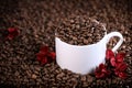 White cup full of coffee beans on a coffee beans background with red chocolate candy and red flowers. Morning espresso. Royalty Free Stock Photo