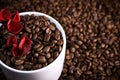 White cup full of coffee beans on a coffee beans background with red chocolate candy and red flowers. Morning espresso. Royalty Free Stock Photo