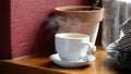 A white cup full of delicious hot coffee or warm tea laying on a table near a window, object closeup, side view, detail, steam