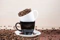 White cup full of roasted coffee beans standing on a black on white plate standing on tablemat.