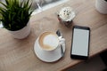 White cup of hot cappuccino on white saucer, black mobile phone, red velvet dessert and green grass in pots on wooden bar table Royalty Free Stock Photo